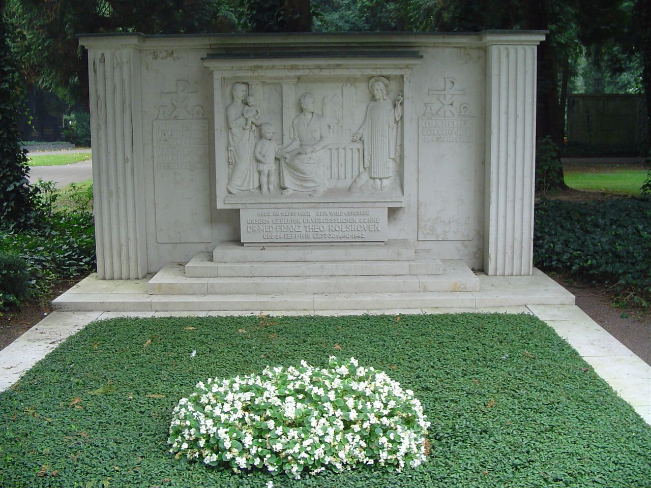 Helle steinerne Grabwand mit zentralem Relief einer Familienszene, flankierenden Pilastern und rundem Beet weißer Blüten auf Nordfriedhof Köln
