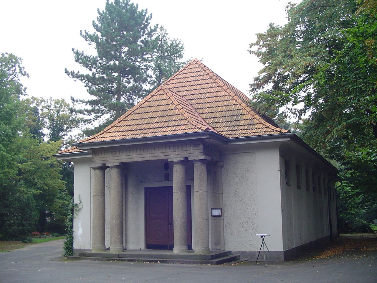 Die kompakte alte Trauerhalle mit hellem Putz, Säulenportal und ziegelgedecktem Walmdach steht inmitten hohen Friedhofsgrüns. auf Nordfriedhof Köln