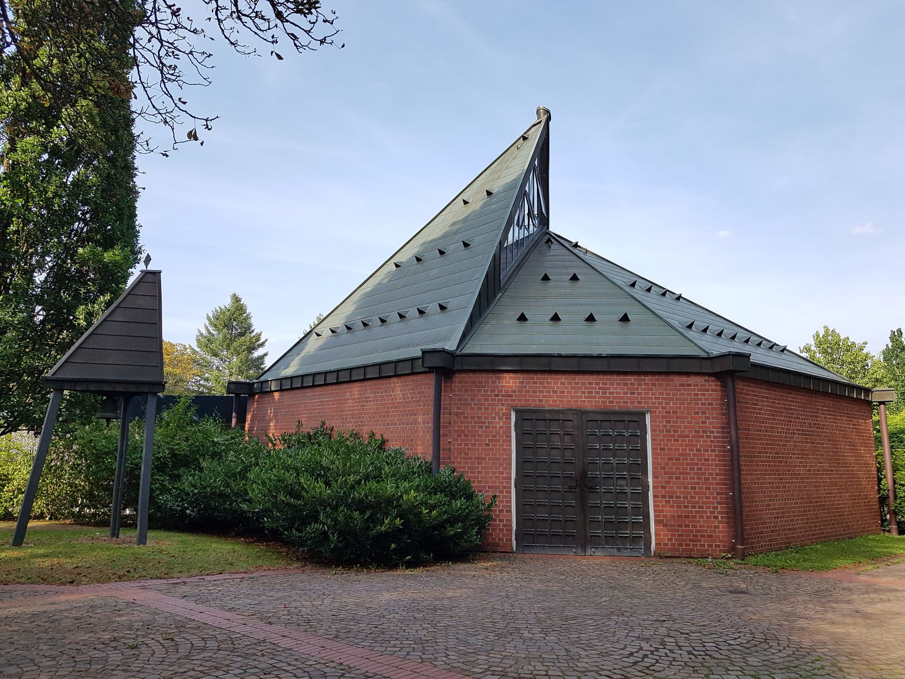 Die moderne Kapelle des Nordfriedhofs als niedriger roter Backsteinbau mit markant gefaltetem grauem Metalldach, dunklen Türen und umgebenden Gehölzen. auf Nordfriedhof Köln