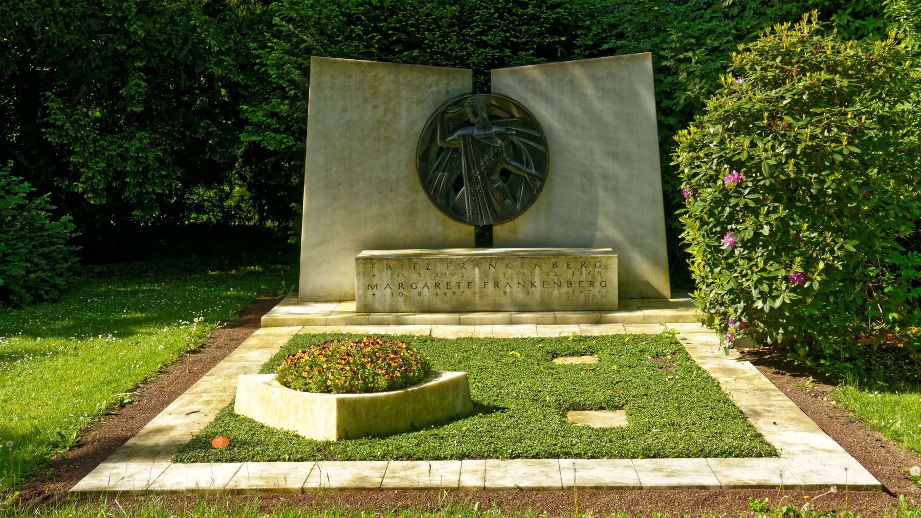 Die gepflegte Grabstätte Frankenberg mit heller, buchartig aufgeklappter Steinwand, rundem Bronzerelief, niedrigem Pflanzbeet und dichtem Grün. auf Nordfriedhof Köln