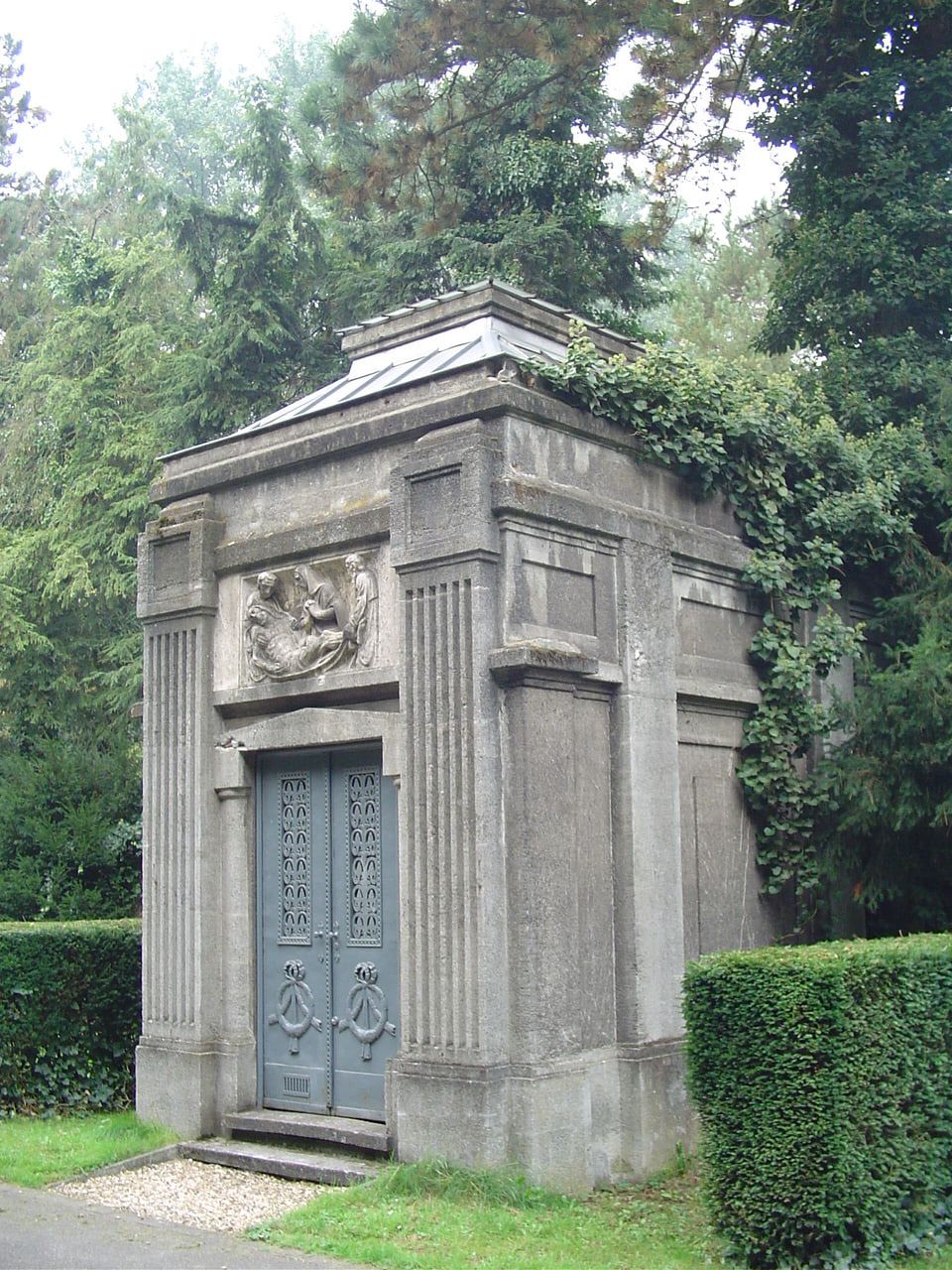 Ein steinernes Jugendstilmausoleum mit reliefgeschmückter Fassade und ornamentierten Metalltüren, flankiert von geschnittenen Hecken und Efeu. auf Nordfriedhof Köln